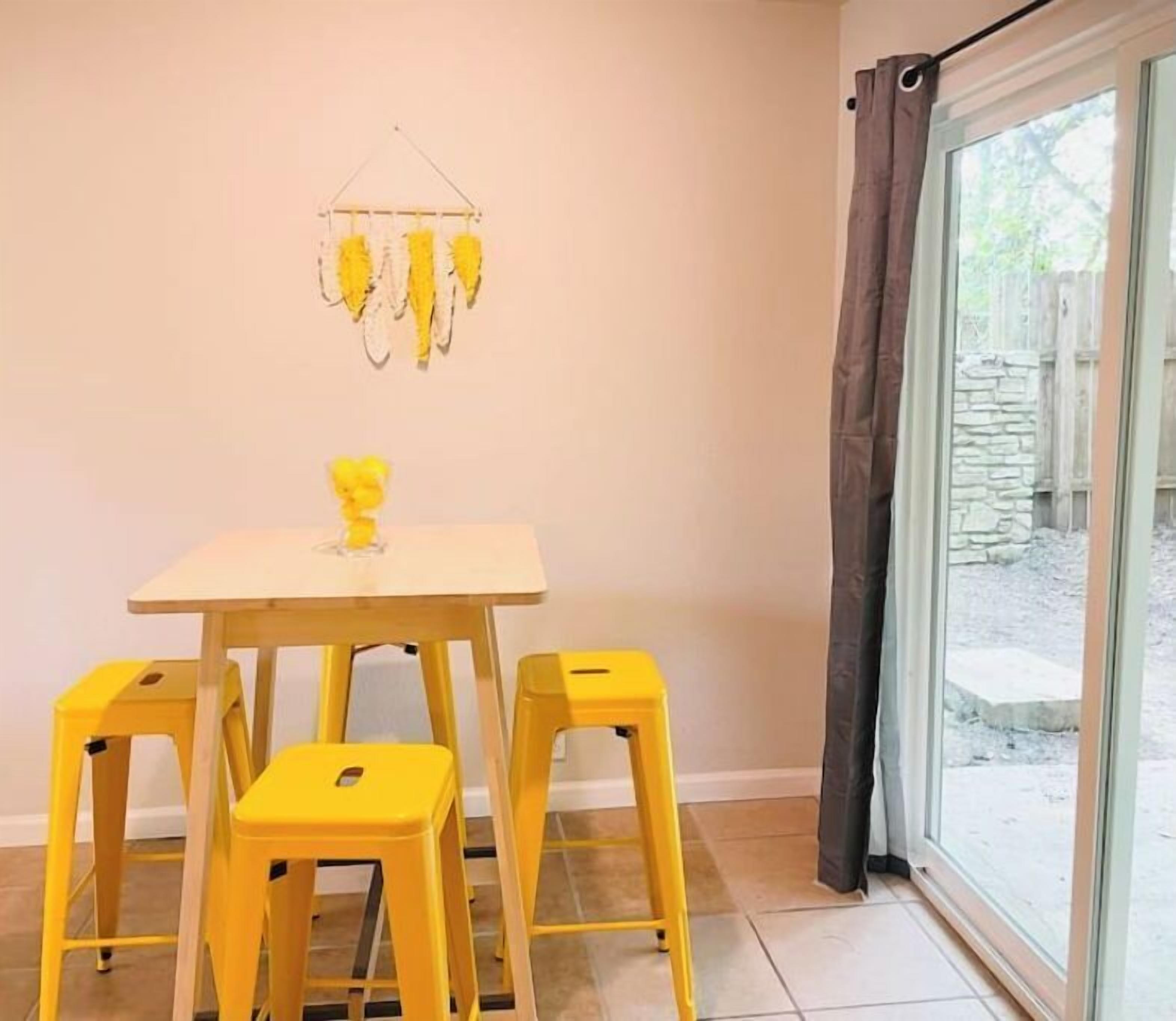 A room with a table and four yellow stools, a hanging decoration, and a view through a window with curtains.