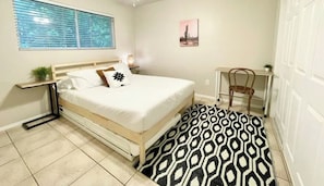 Een slaapkamer met een bed, bureau, stoel, raam met zonwering en een foto van een cactus aan de muur.