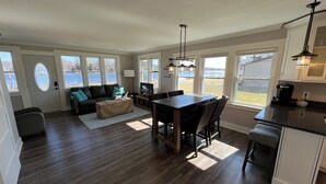A spacious living room with a dark wood floor, a black leather sofa, a coffee table, and a large window overlooking a body of water. - Perfect Lake House Getaway! (Waterford)