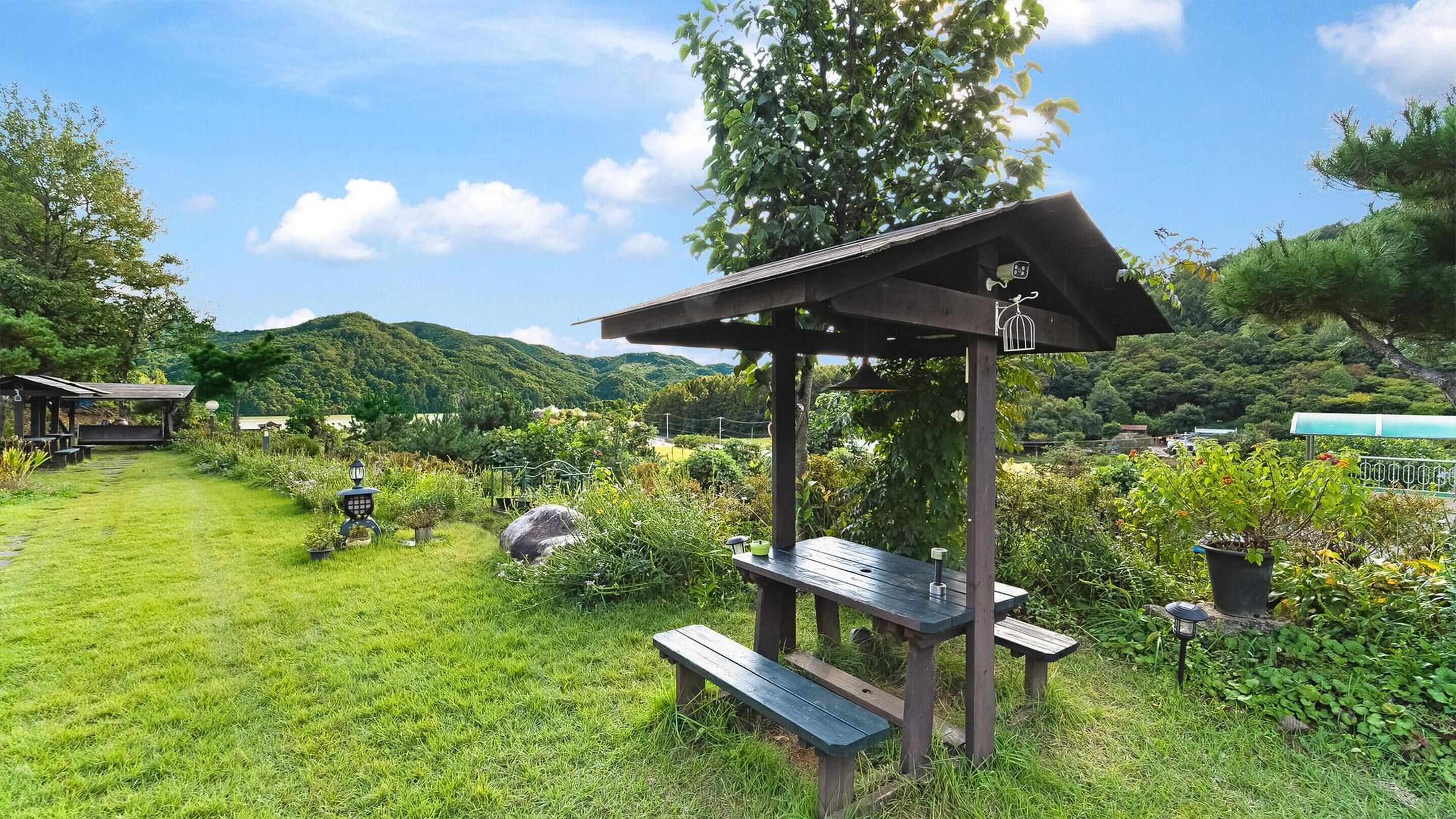 Een houten tuinpaviljoen met een schuin dak en een hangende vogelkooi, omgeven door weelderige begroeiing en een picknicktafel.
