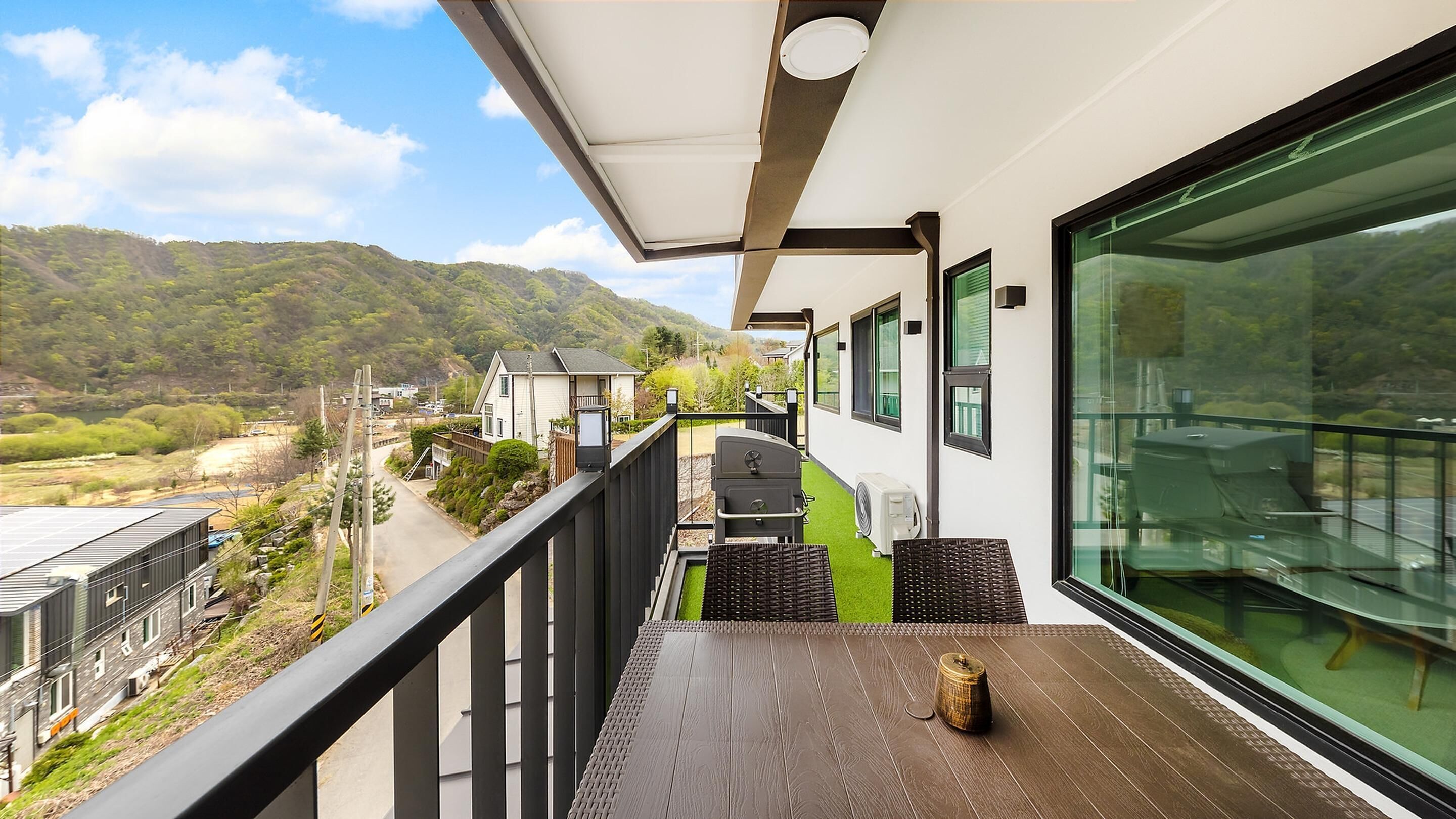 A balcony with a wooden table, a grill, and a view of a green landscape and mountains.