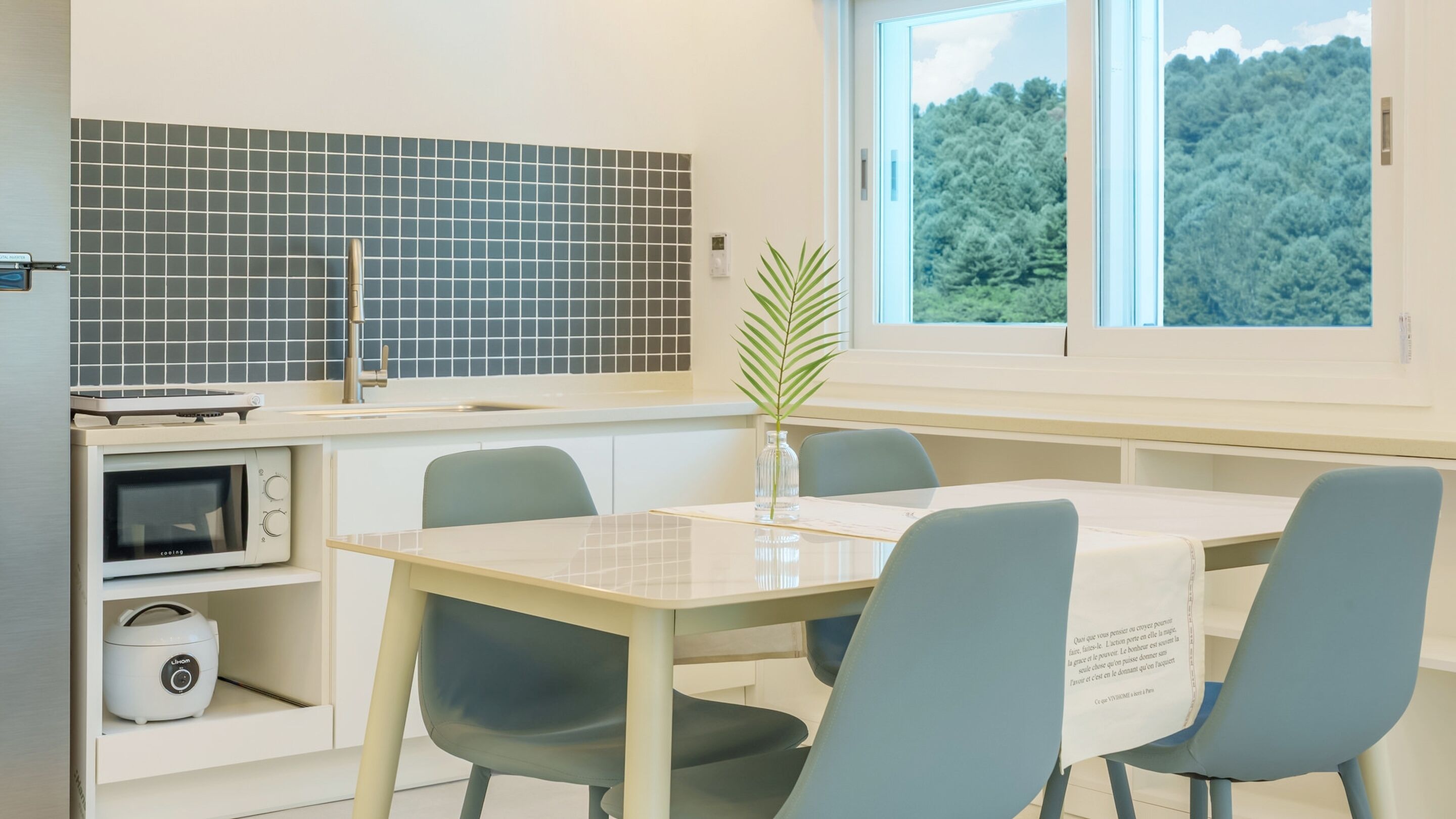 A modern kitchen with a microwave, oven, and a sink. There's a dining table with chairs and a window with a view of trees.