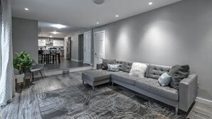 Ein modernes Wohnzimmer mit einem grauen Ecksofa, einem gemusterten Teppich und einem Essbereich im Hintergrund.