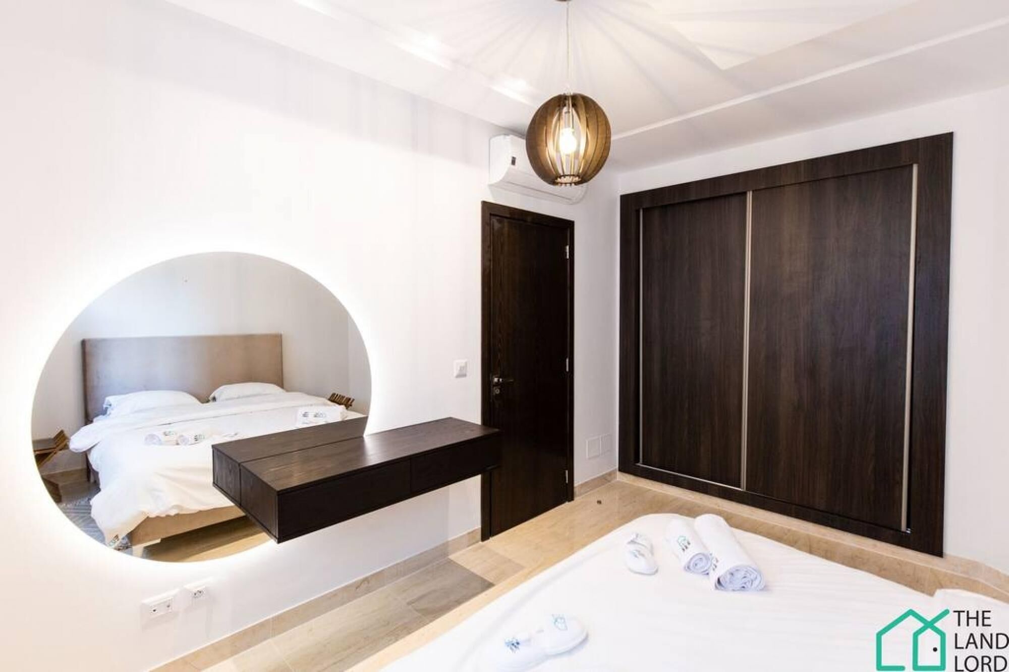 A modern bedroom with a bed, a circular mirror, a dark wooden wardrobe, and a hanging light fixture.