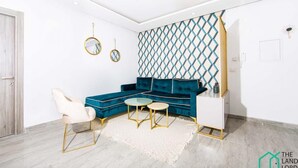A modern living room with a blue sectional sofa, a round coffee table, and a white side table. There are two gold-framed mirrors on the wall, a patterned wall divider, and a white door. - New 1bd With Swimming Pool 5mn to Lac Carthage (Tunis)