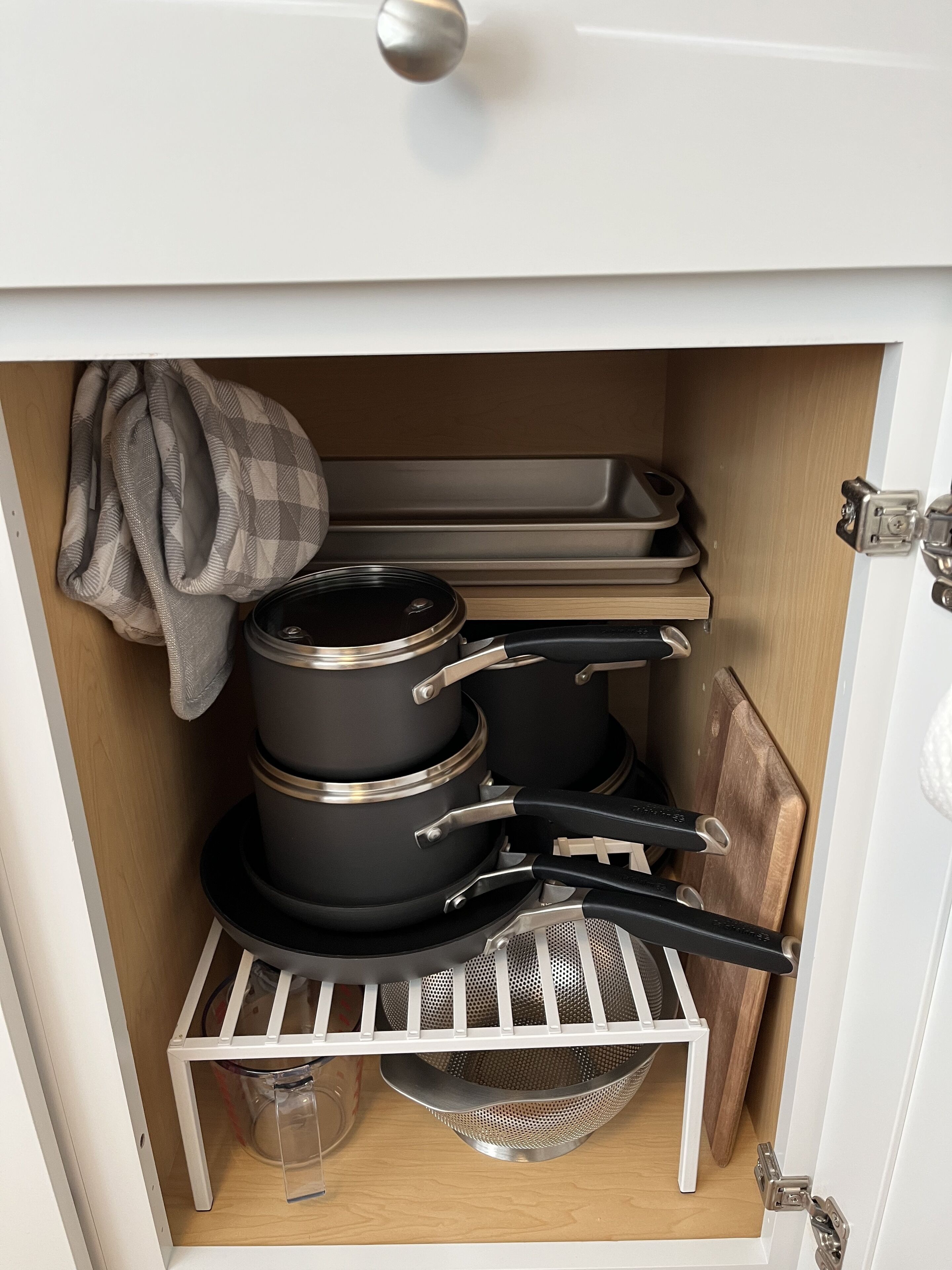Um armário de cozinha com panelas, frigideiras, um escorredor e uma tábua de corte.