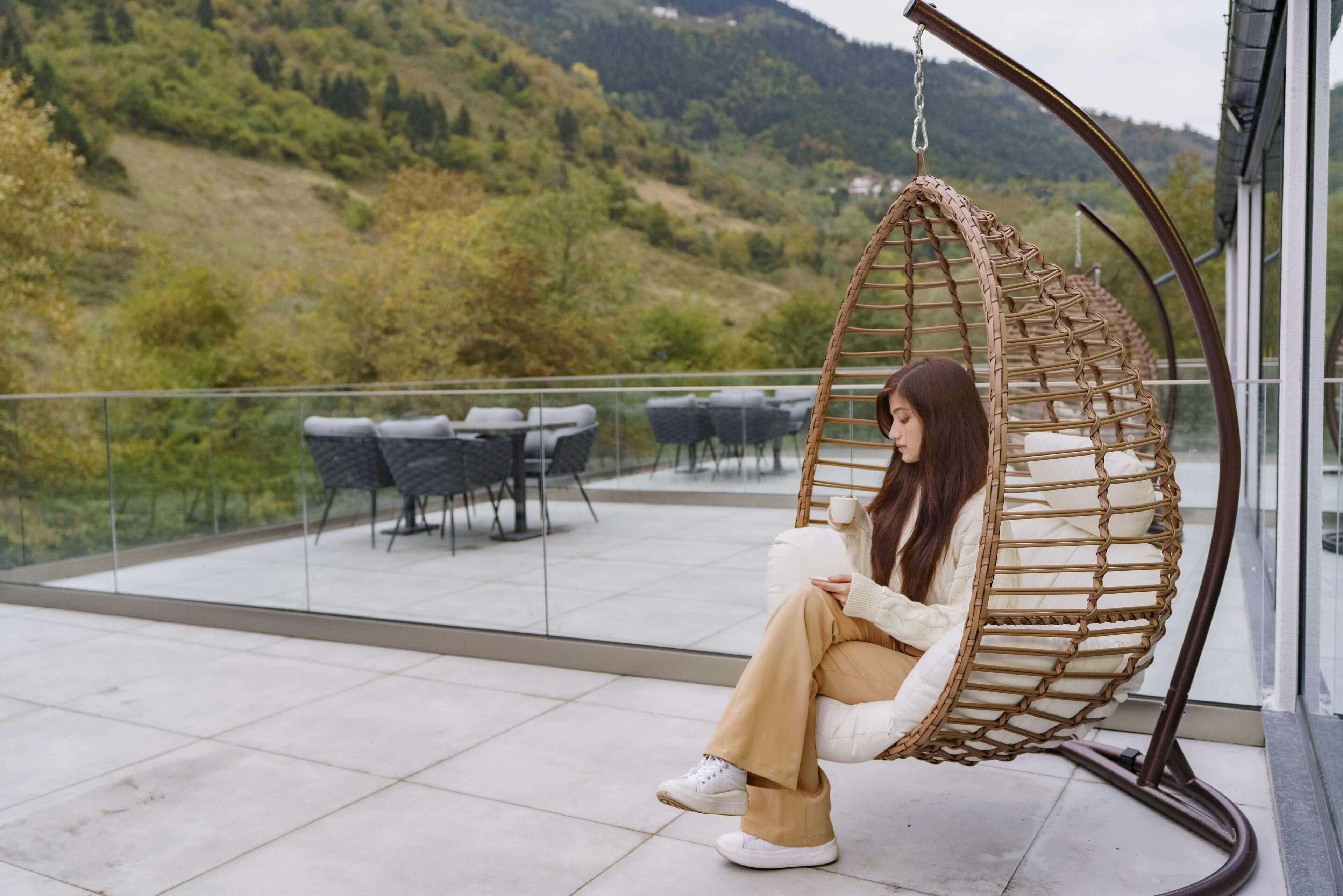 Una persona sentada en una silla colgante de mimbre en un balcón, con vista a un paisaje montañoso.