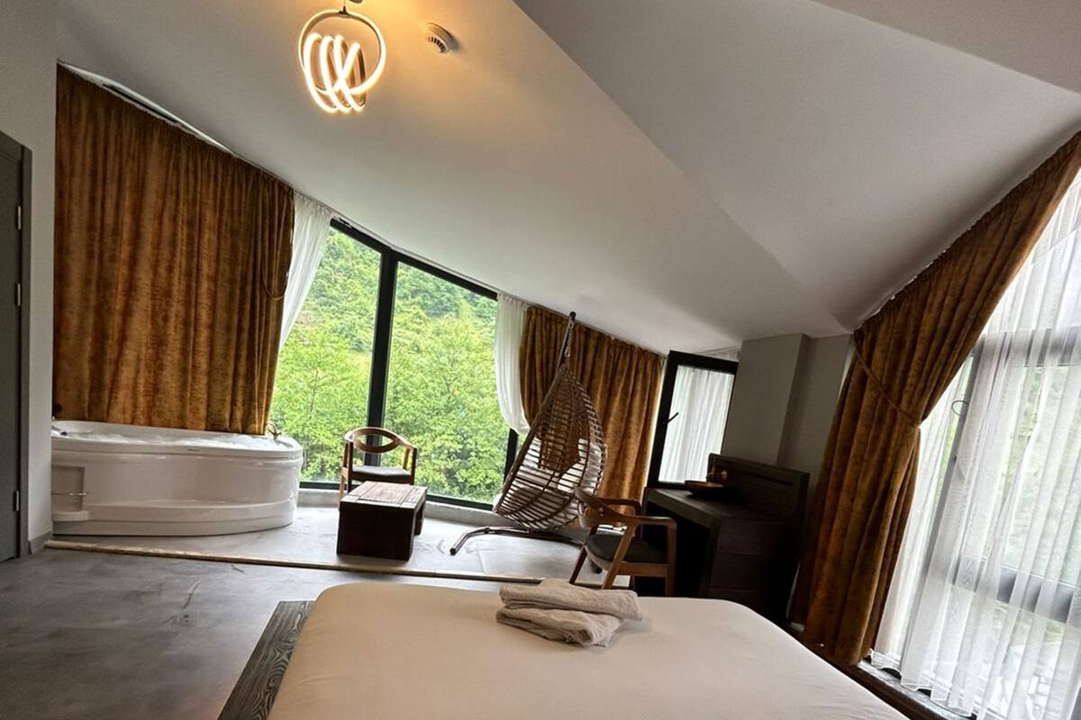 A modern bedroom with a large bed, a hanging chair, a bathtub, and a view of greenery outside.