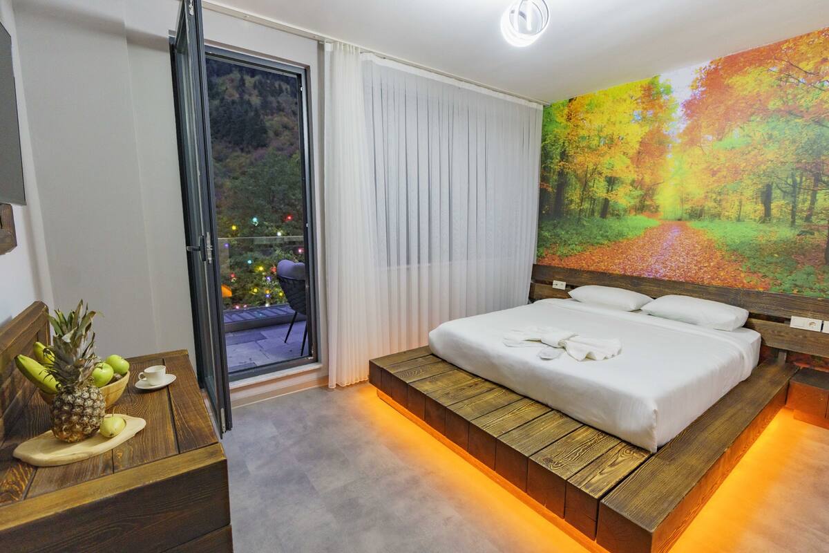 A modern hotel room with a large bed, a wooden bench with fruit, a view of a forest through a window, and a cityscape at night.