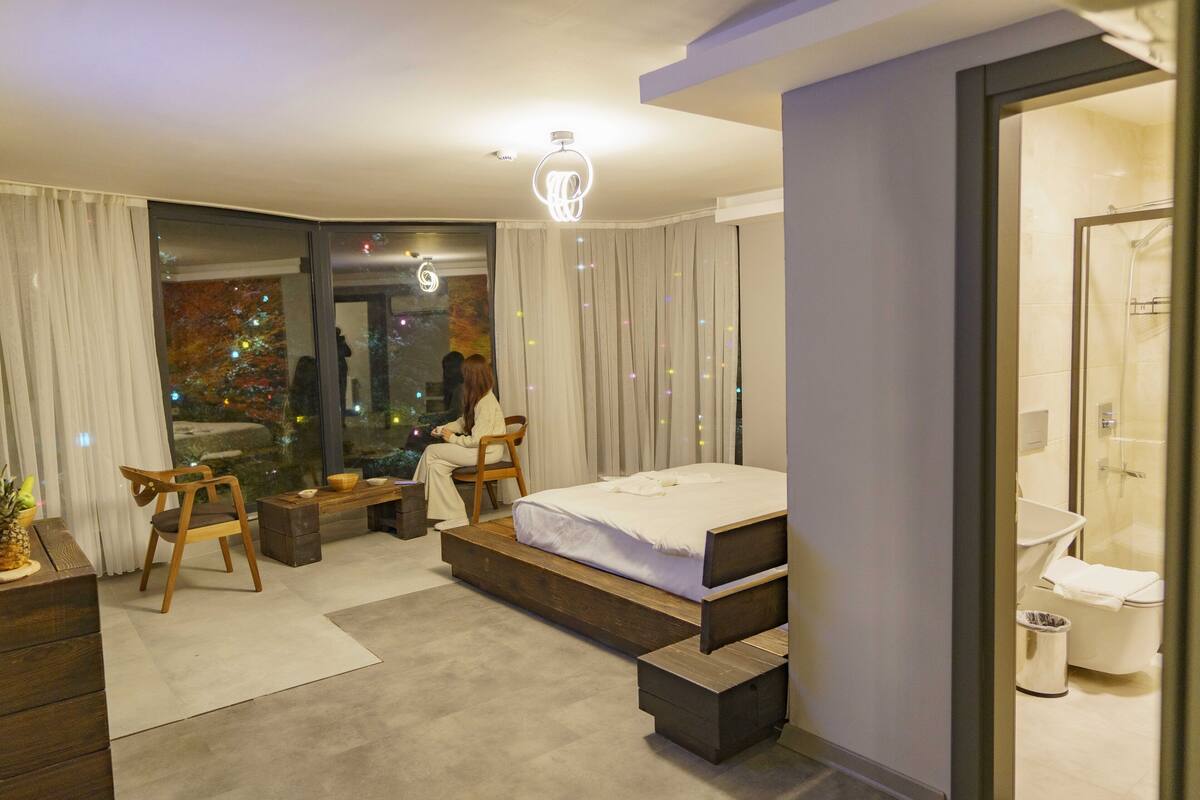 A modern bedroom with a bed, a desk, and a chair. There is a window with curtains and a view of a cityscape.
