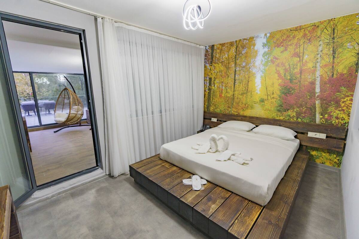 A bedroom with a bed, a wooden platform, a large window with curtains, and a view of an outdoor seating area.