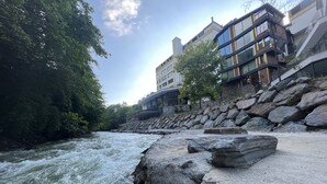 A river with a rocky riverbank, a stone wall, and modern buildings with large windows. - MacarTaşı Hotel  (Maçka)