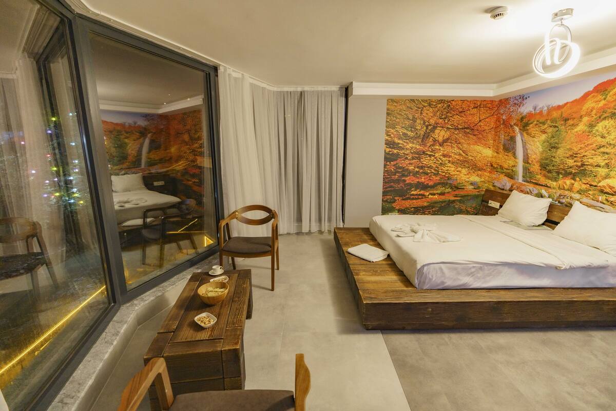 A modern hotel room with a large bed, a dining table, and a view of a forest through a glass wall.