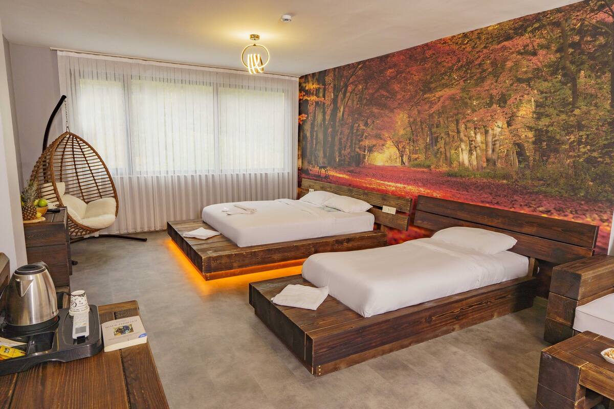 A modern hotel room with two wooden beds, a hanging chair, a large forest-themed wall mural, and a bedside table with a kettle and a cup.