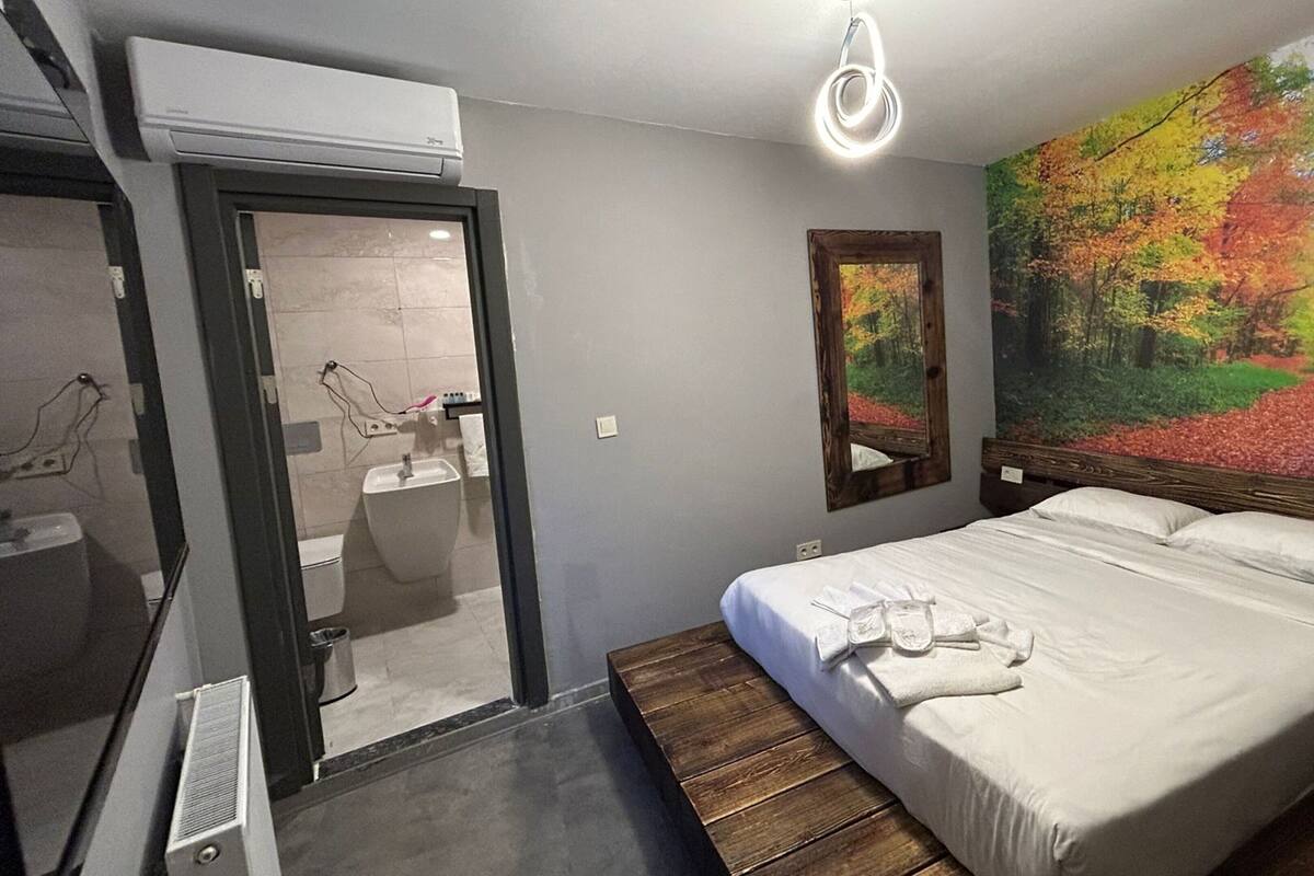 A bedroom with a bed, a large mirror, a painting of a forest, and a bathroom visible through an open door.
