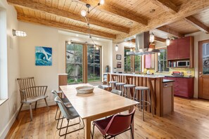A wooden dining table with chairs, a kitchen area with a microwave, and a large window with a view of trees.