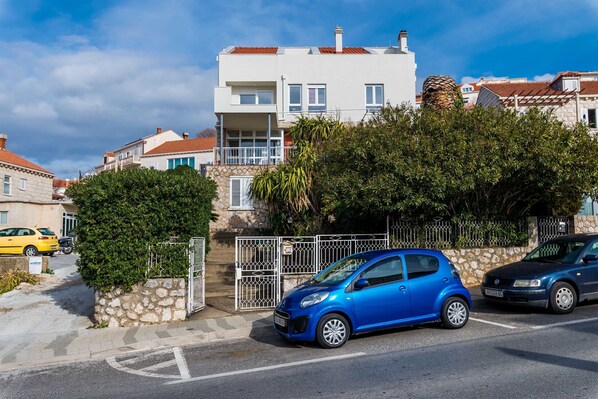 A residential street with a blue car parked in front of a white building with a stone wall and a metal gate. - Bovolini's Room (Dubrovnik)