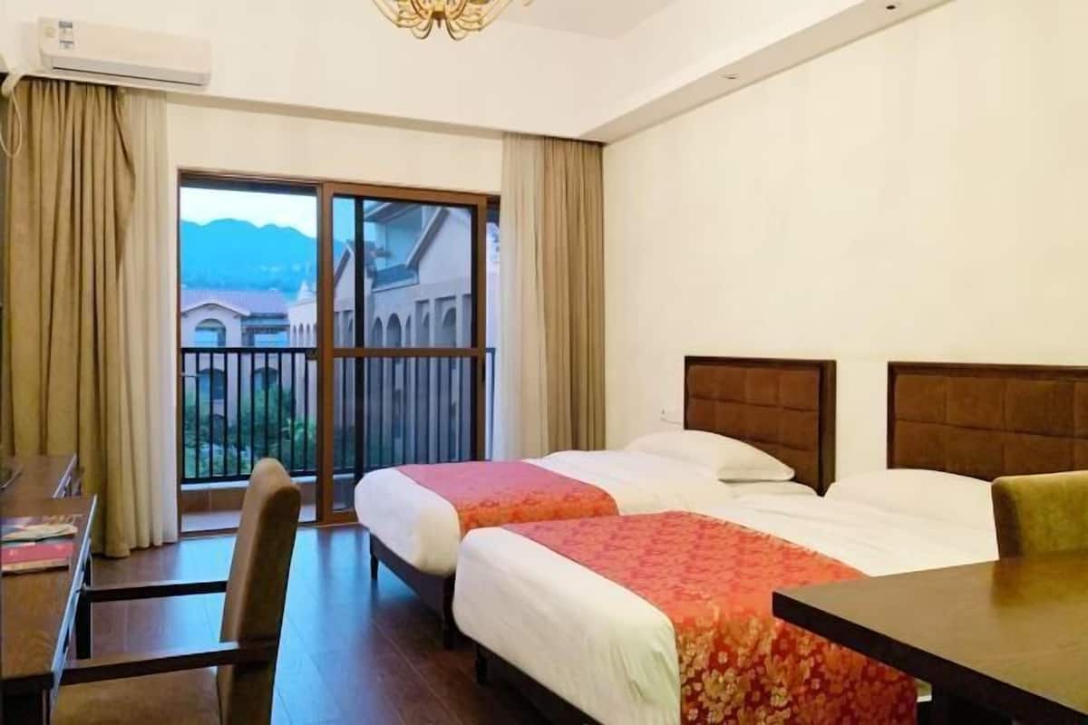 A hotel room with two beds, a desk, and a chair. There is a large window with a view of buildings and mountains.