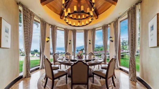 Mesa de jantar redonda com cadeiras, um candelabro e grandes janelas com cortinas.