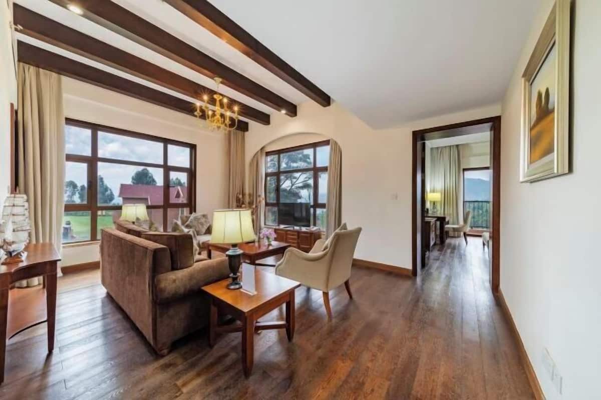 A room with wooden flooring, a sofa, a table, a lamp, and large windows with a view of trees and buildings.