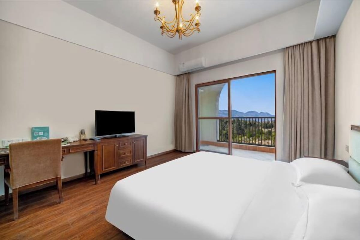 Una habitación de hotel con una cama grande, un escritorio con televisor, una silla y vistas a montañas a través de un ventanal amplio.
