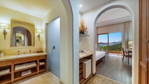 Casa de banho de hotel com um espelho grande, duas pias e zona de duche. A casa de banho dá para um quarto com uma janela ampla e vista panorâmica.