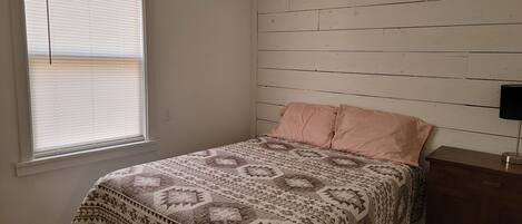 A bedroom with a bed, a window with blinds, a wooden wall, and a nightstand with a lamp.