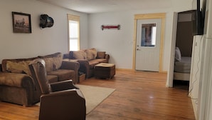 Ein Wohnzimmer mit einem braunen Ledersofa, einem weißen Sessel und einem Holztisch. Eine Tür führt in einen anderen Raum, und an der Wand ist ein Fernseher befestigt.