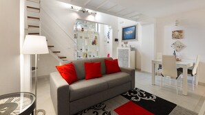 Un salon moderne avec un canapé gris, des coussins rouges, une table à manger avec des chaises, un escalier et une bibliothèque.