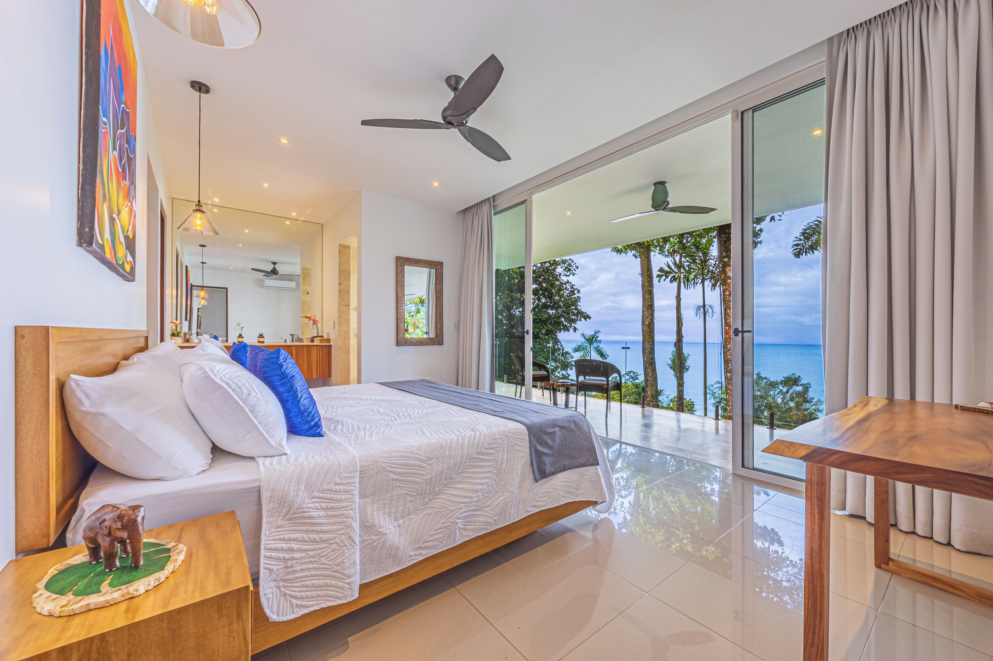 Ein modernes Hotelzimmer mit einem Bett, einem Nachttisch aus Holz, einem Deckenventilator, einem großen Fenster mit Blick aufs Meer und einem Essbereich mit Tisch und Stühlen.