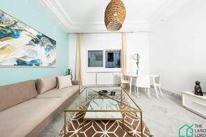 Un salon moderne comprenant un canapé, une table basse en verre, un coin repas avec une table et des chaises blanches, et un grand tableau abstrait accroché au mur.