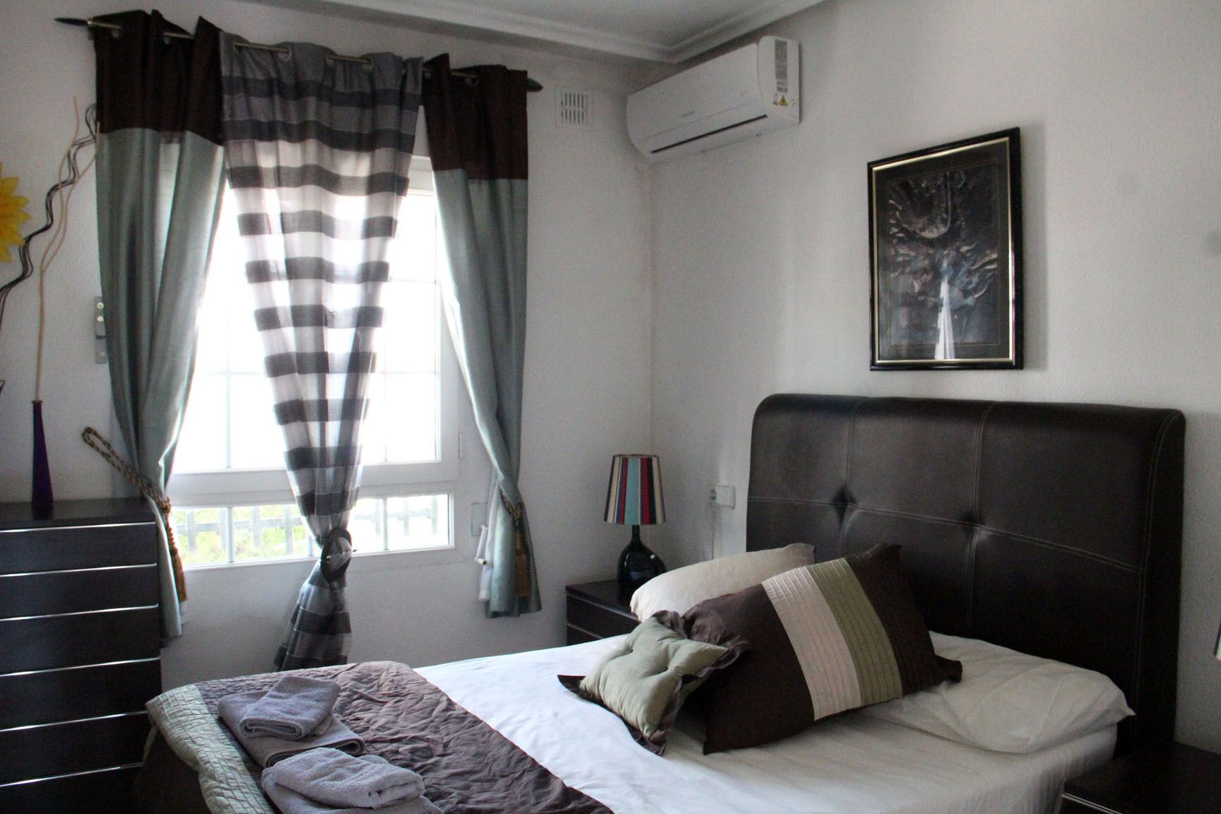 A bedroom with a bed, a nightstand, a lamp, a window with curtains, and an air conditioning unit.