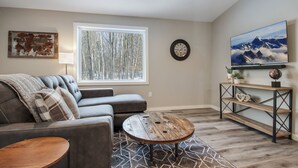 A living room with a gray sectional sofa, a round coffee table, a large window with a view of trees, a mounted TV, and a wall clock. - Up North Living!  Fully renovated one-level living home in wooded lot near lake. (Cumberland)