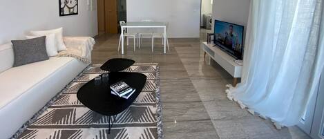 A modern living room with a sofa, coffee table, and TV. There is a dining area with a table and chairs.