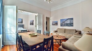 A spacious living room with a wooden dining table, a sofa, and a coffee table. There are two doors leading to other rooms, and the walls are adorned with framed pictures. - Elegant Apartment in Parliament Square (Athens)