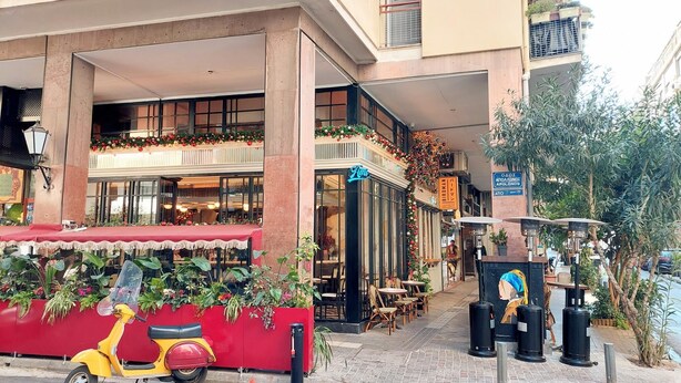 A street view with a restaurant named 'Ami' featuring outdoor seating, a yellow scooter parked in front, and a sign for 'Birdman' across the street. - Elegant Apartment in Parliament Square (Athens)