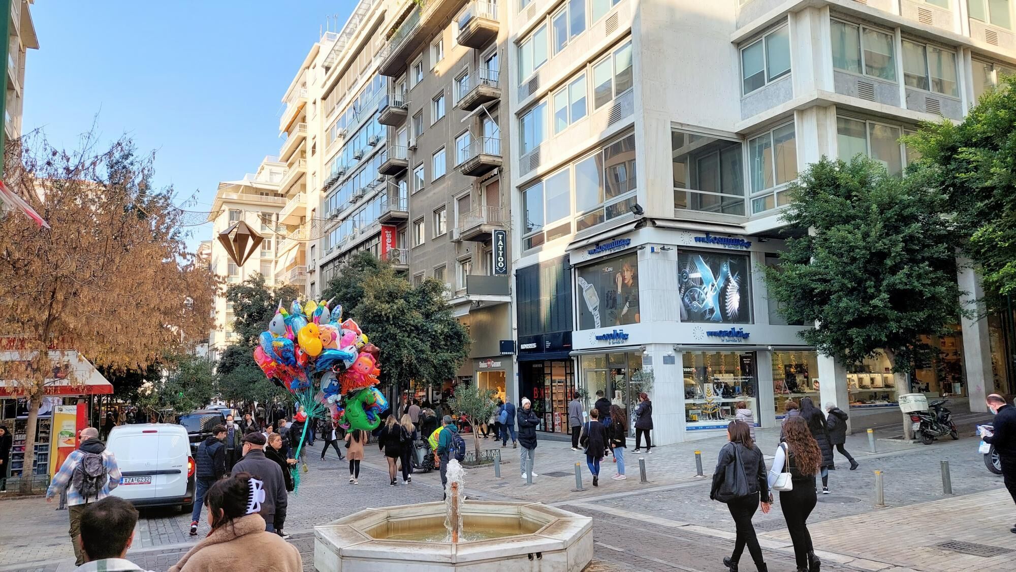 Eine belebte Stadtstraße mit Fußgängern, einem Brunnen und verschiedenen Geschäften.