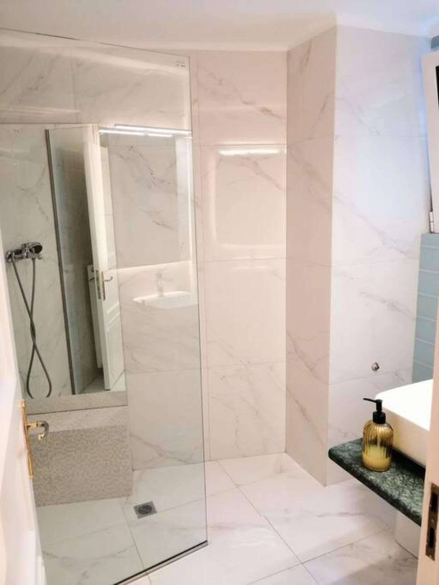 A bathroom with a glass shower enclosure, a marble-tiled floor, and a small sink with a gold soap dispenser.