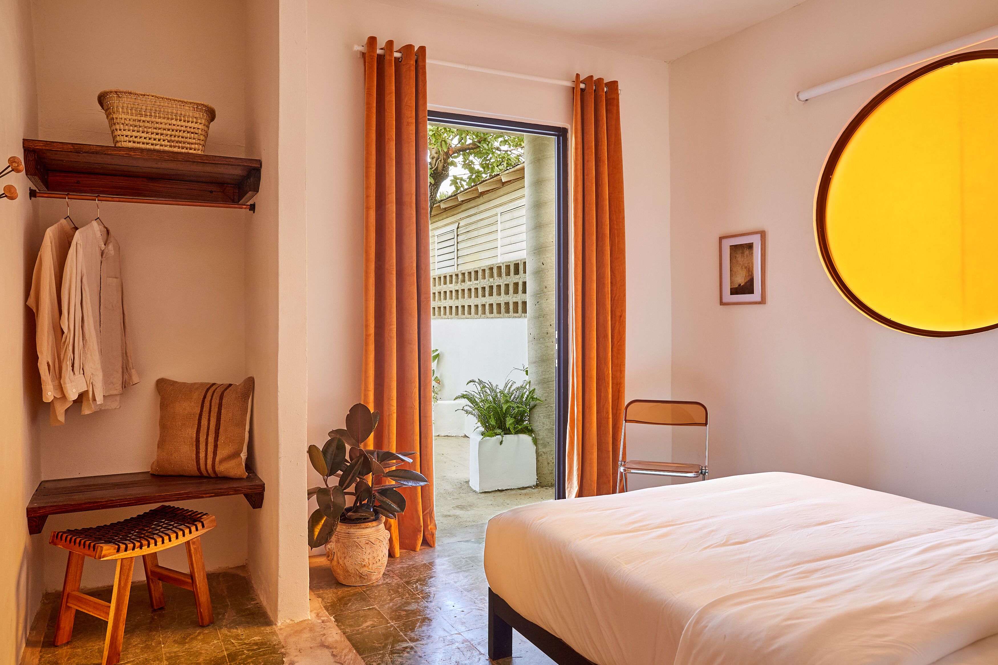 A bedroom with a bed, a chair, a bench, a shelf with clothes, a basket, and a potted plant.