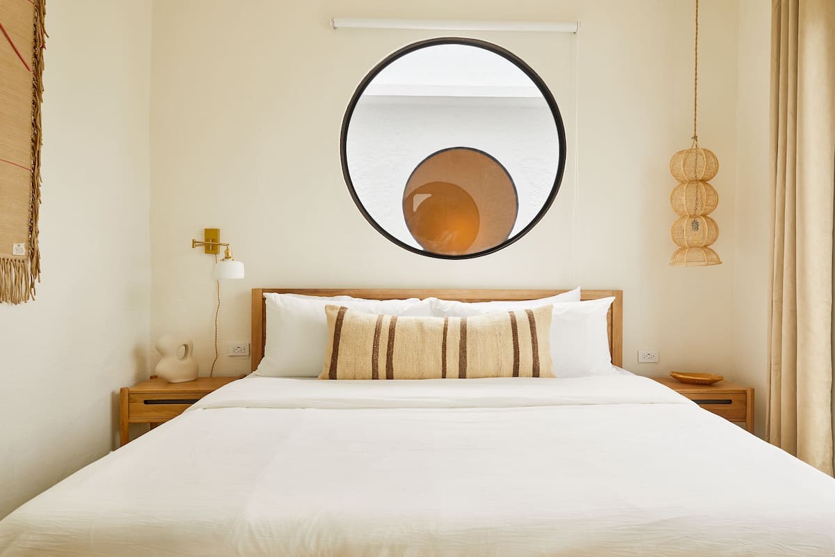 A neatly made bed with white linens and striped pillows, a round mirror on the wall, and a bedside lamp.