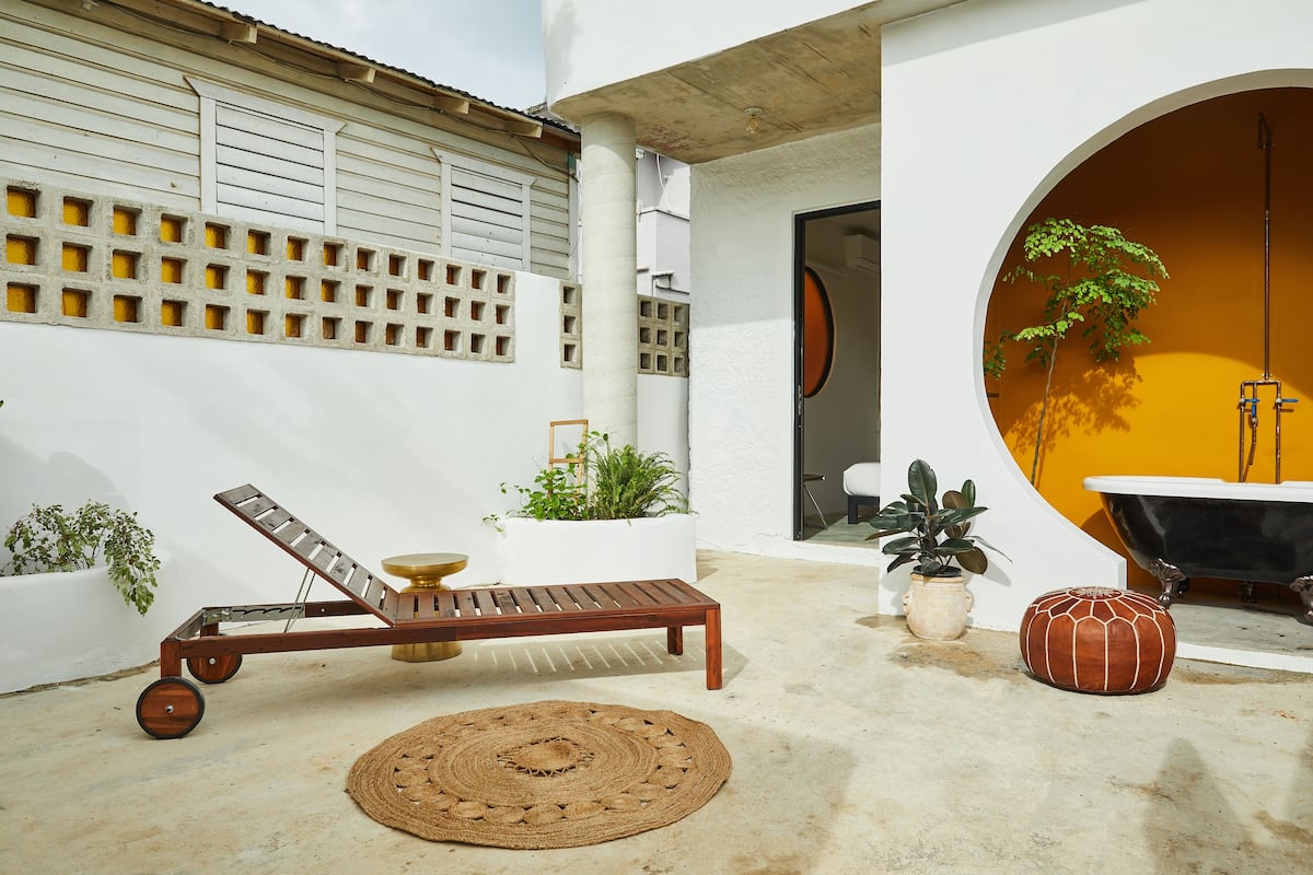 A sunlit outdoor patio with a wooden lounge chair, a woven rug, potted plants, and a bathtub.
