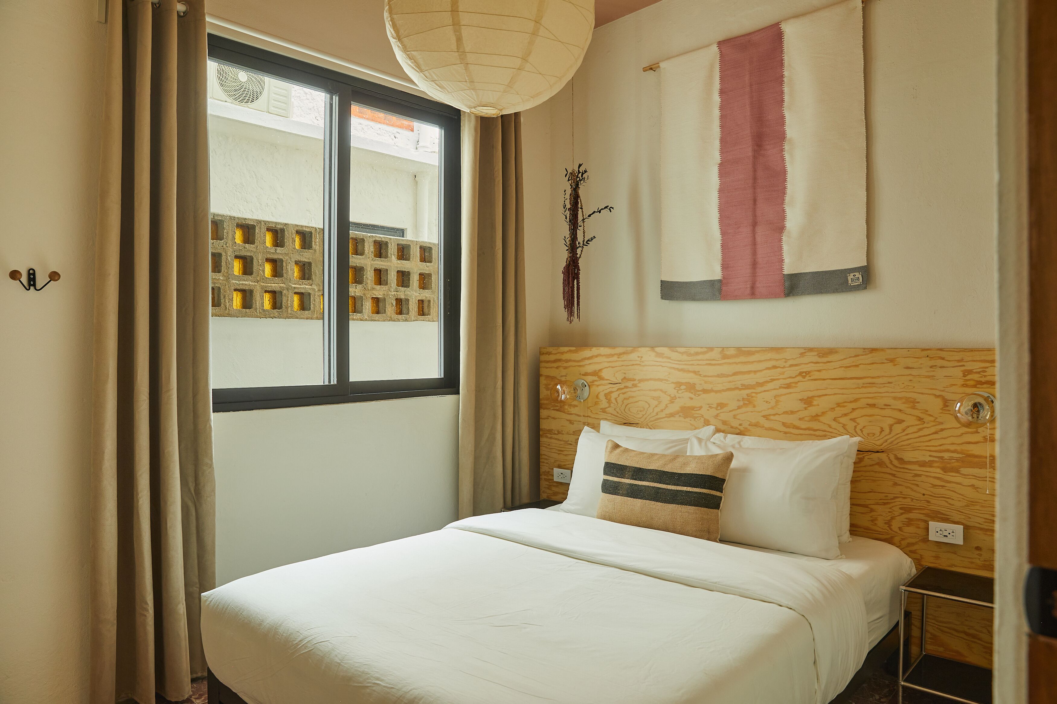 Ett hotellrum med en sänggavel i trä, ett stort fönster med persienner, en säng med vita sängkläder och en väggmonterad lampa.
