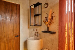 A bathroom with a wooden door, a white sink, a mirror, and a shelf with dried flowers. - APT by DW (San Juan)