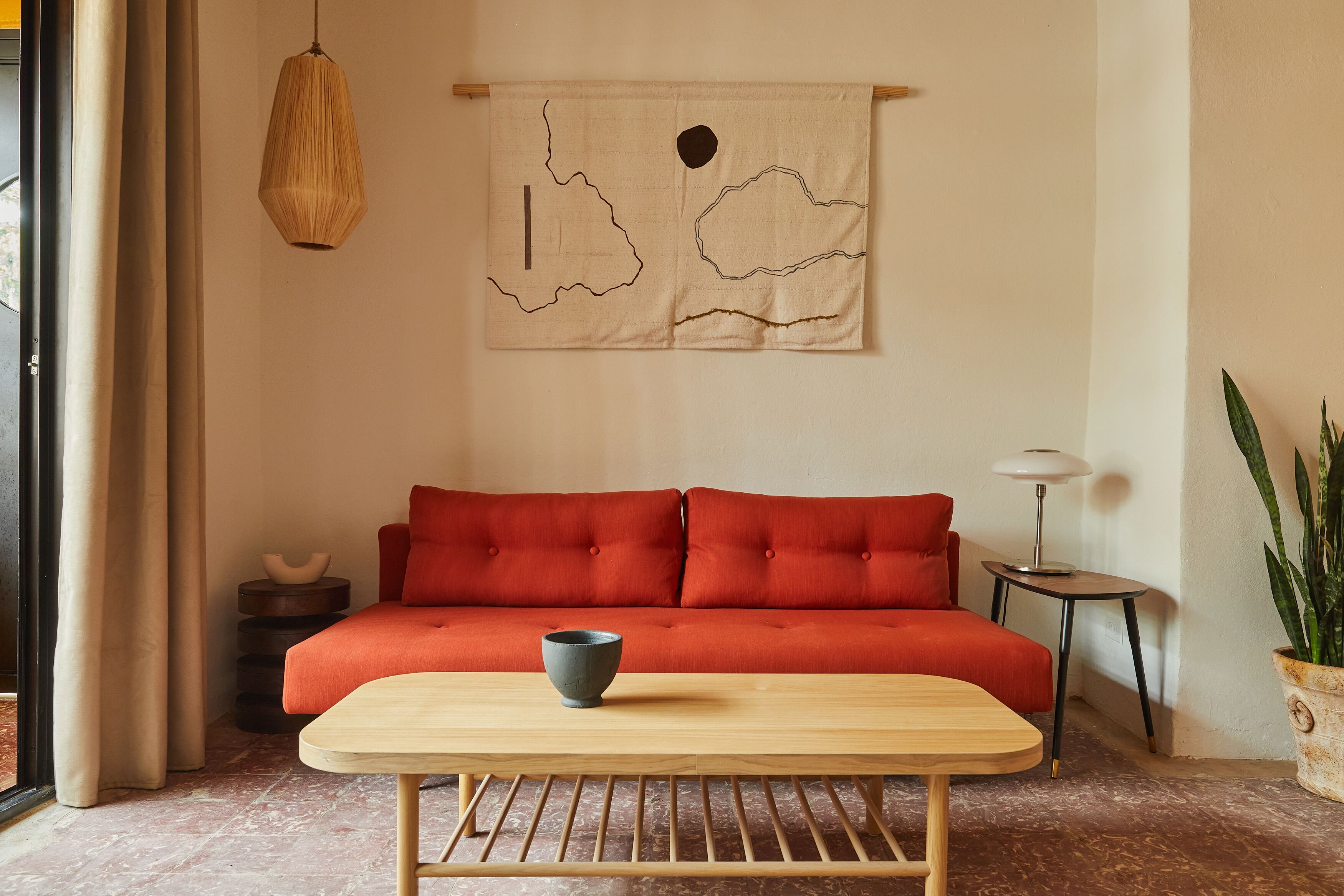 A room with a red sofa, a wooden table with a bowl, a hanging light, and a wall tapestry.