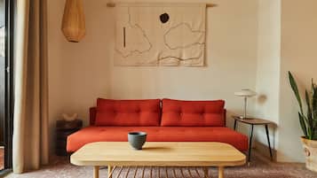 A room with a red sofa, a wooden table with a bowl, a hanging light, and a wall tapestry.