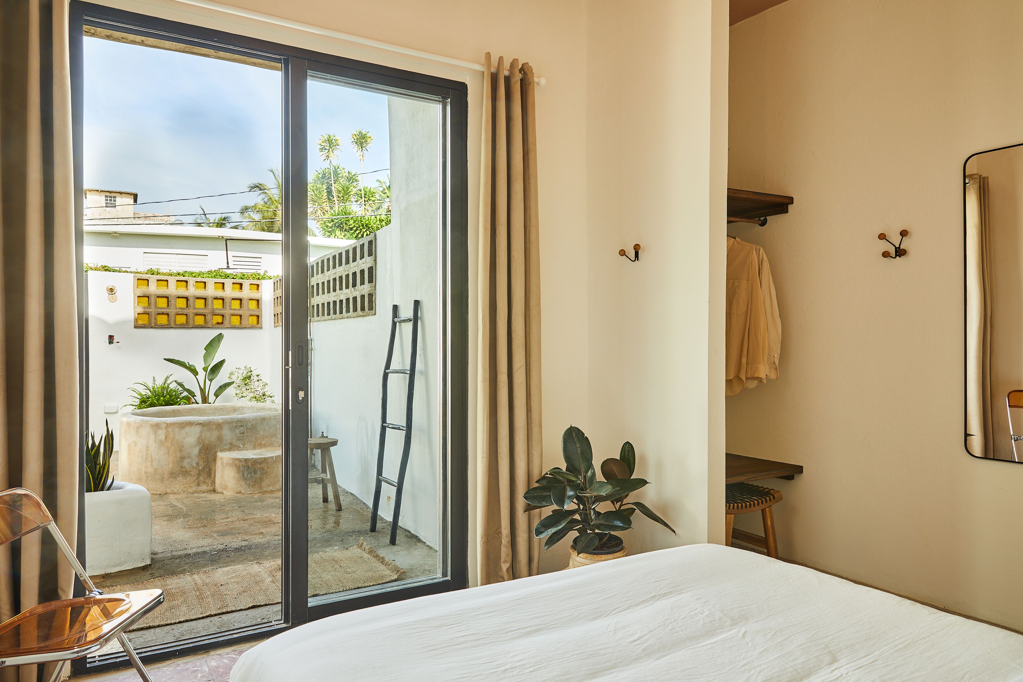A hotel room with a sliding glass door leading to an outdoor area, a bed, a chair, a small table, and a potted plant.
