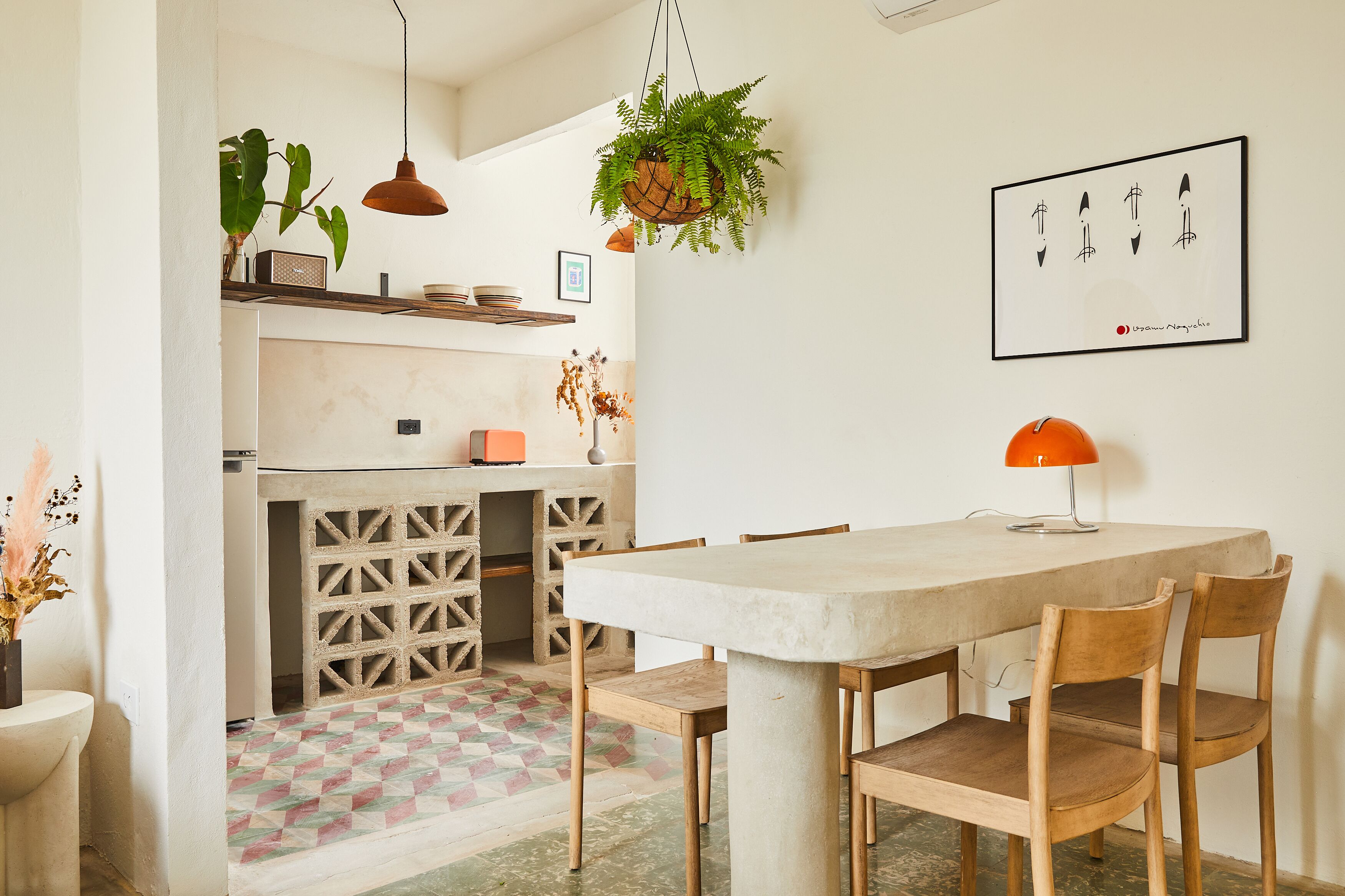 Un comedor con una mesa grande, sillas de madera, una planta colgante, una lámpara colgante y una obra de arte enmarcada en la pared.