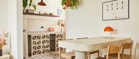 Un comedor con una mesa grande, sillas de madera, una planta colgante, una lámpara colgante y una obra de arte enmarcada en la pared.