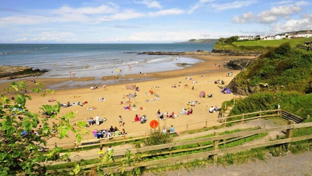 Une plage où des gens se prélassent au soleil et se baignent, une clôture en bois et une falaise herbeuse.