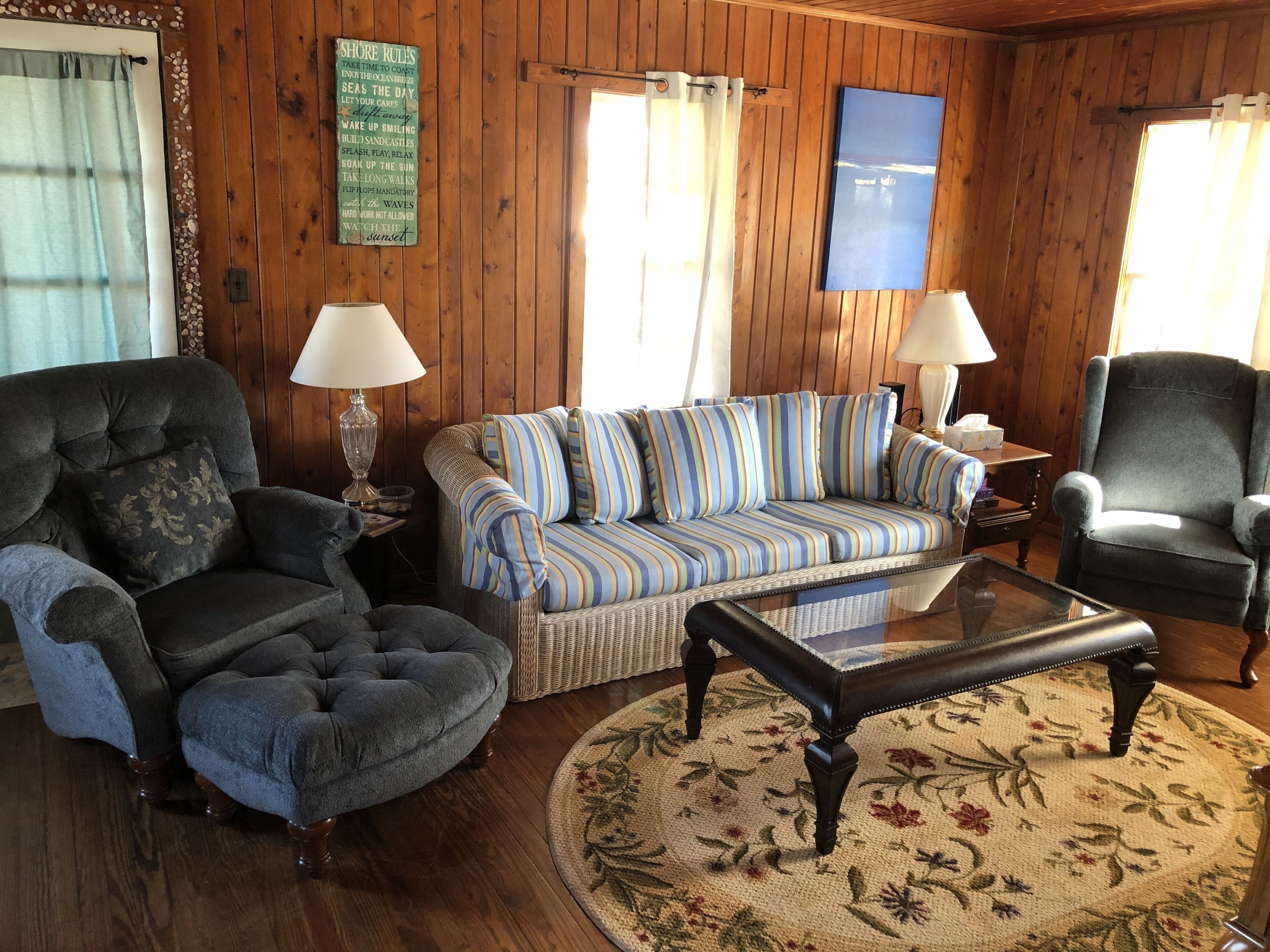 A cozy living room with wooden walls, a striped sofa, two armchairs, a glass coffee table, and a floral rug.